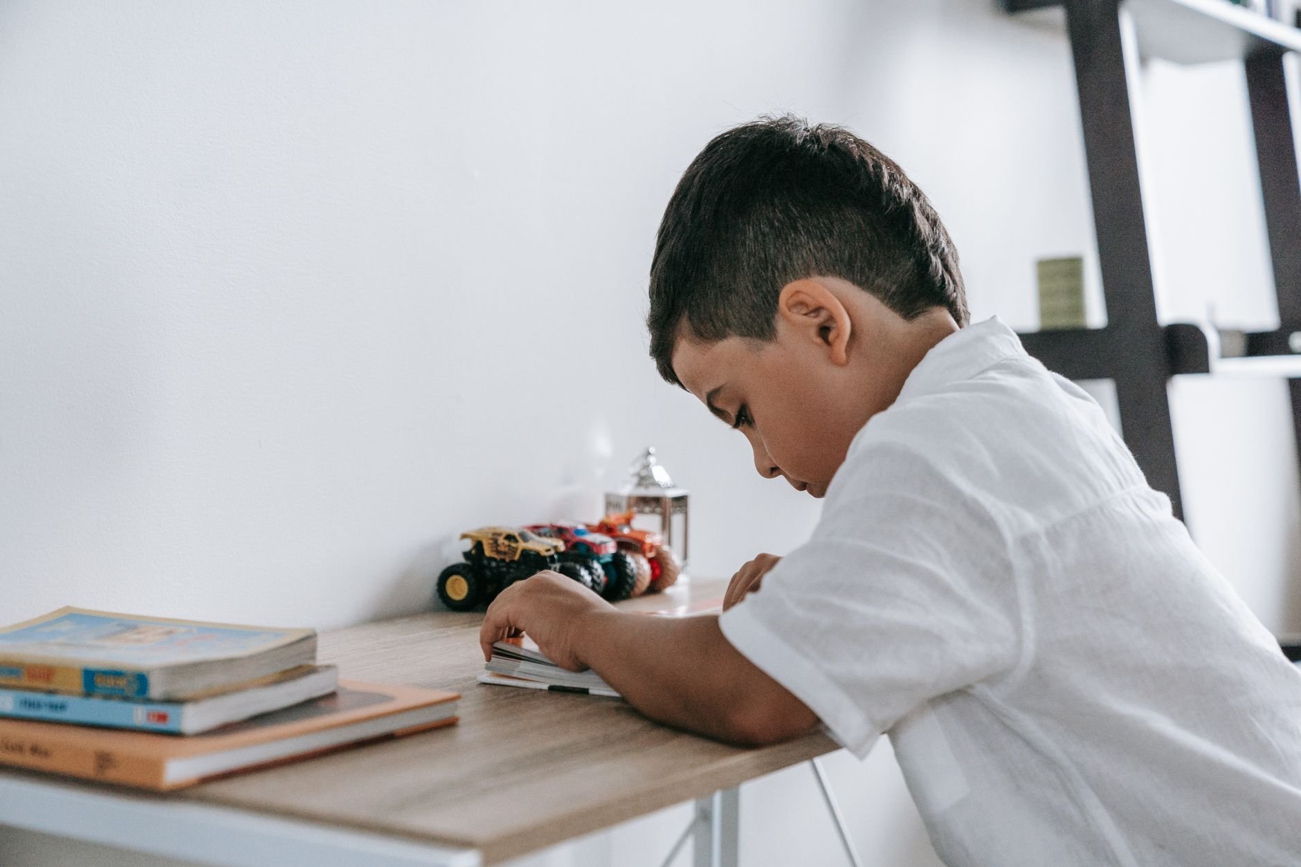 Child engrossed in reading at a desk with toy trucks in a bright room.