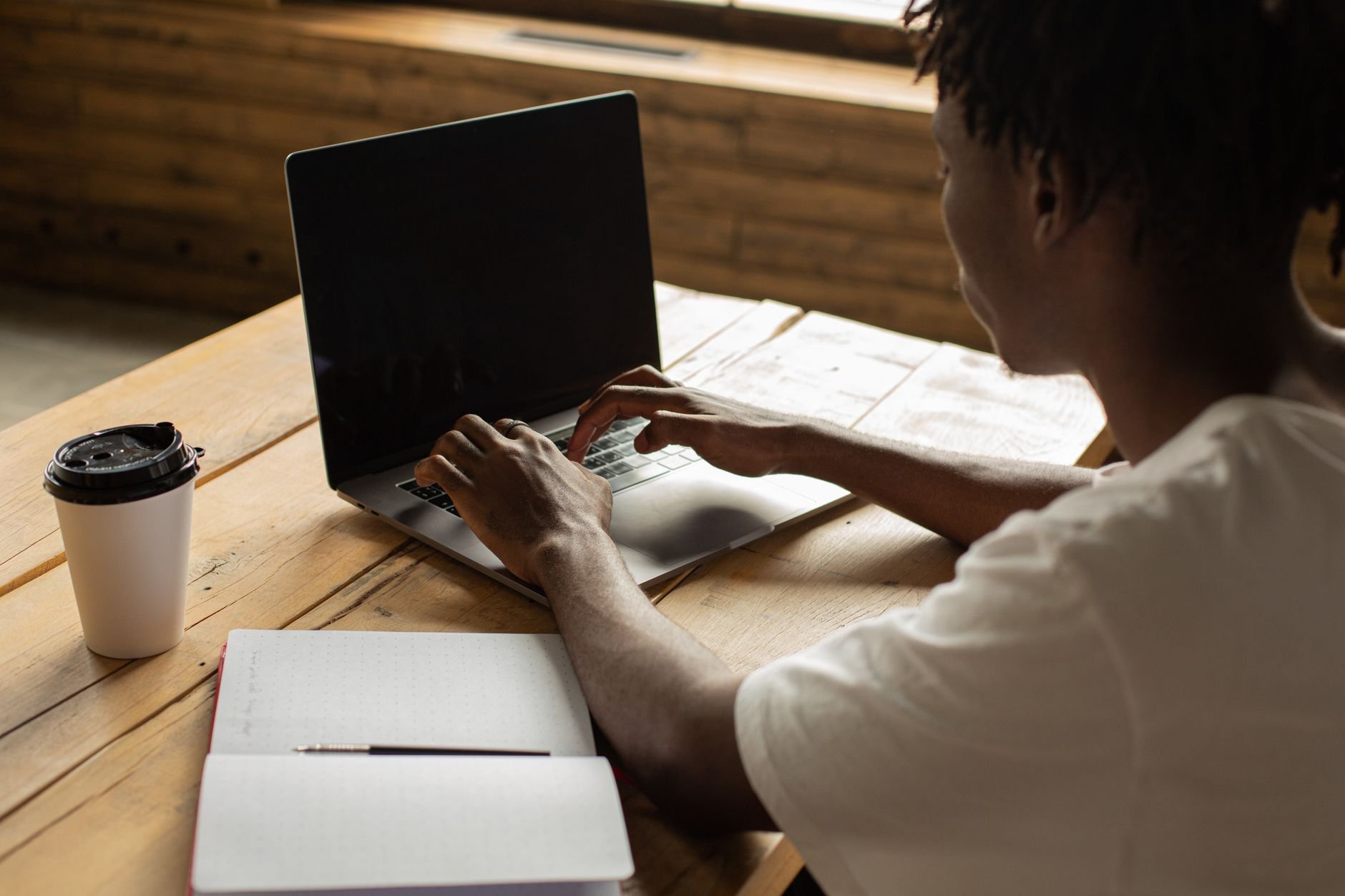 Young man working on a laptop in a cozy cafe with coffee and notebook on table.